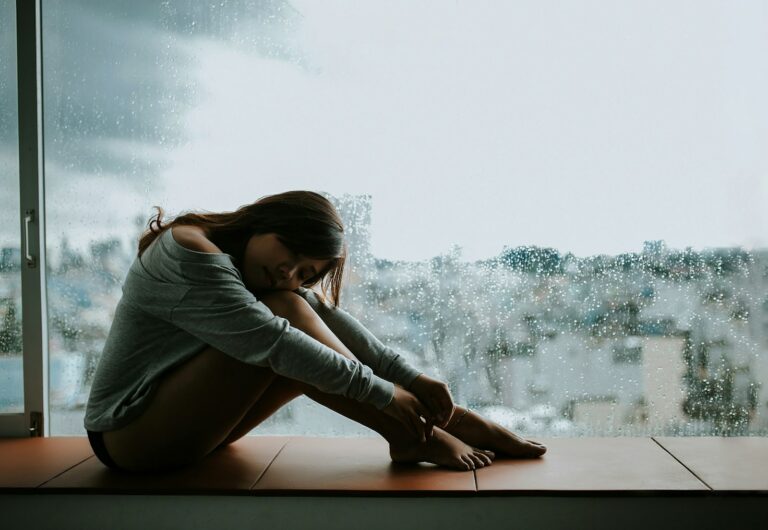 Mujer sentada junto a una ventana con lluvia, abrazada a sus rodillas, sin poder dejar de pensar en él